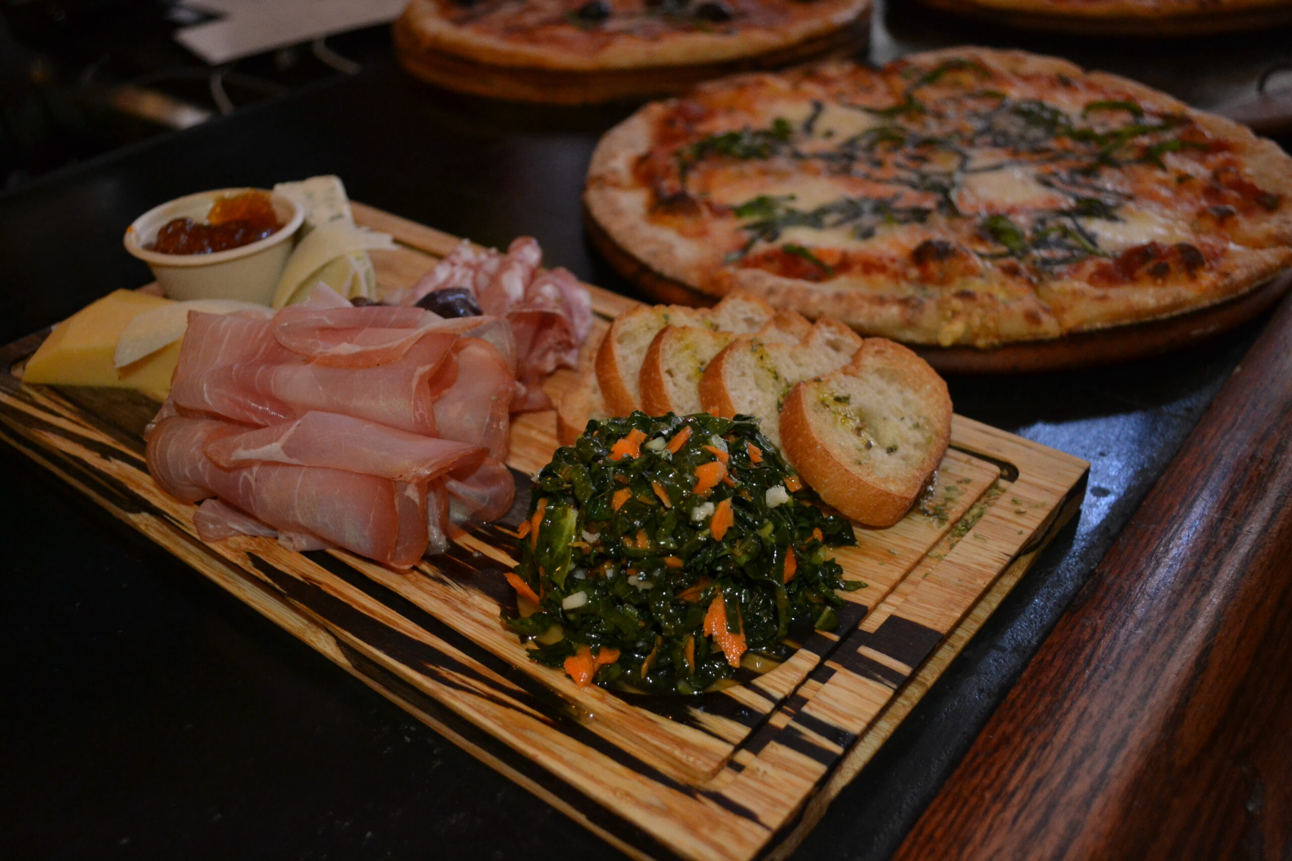 A rustic wooden charcuterie board with prosciutto, artisan cheeses, garlic crostini and kale salad beside wood-fired margherita pizza at a Bay Area restaurant, the kind of shareable spread that anchors a seasonal social media campaign
