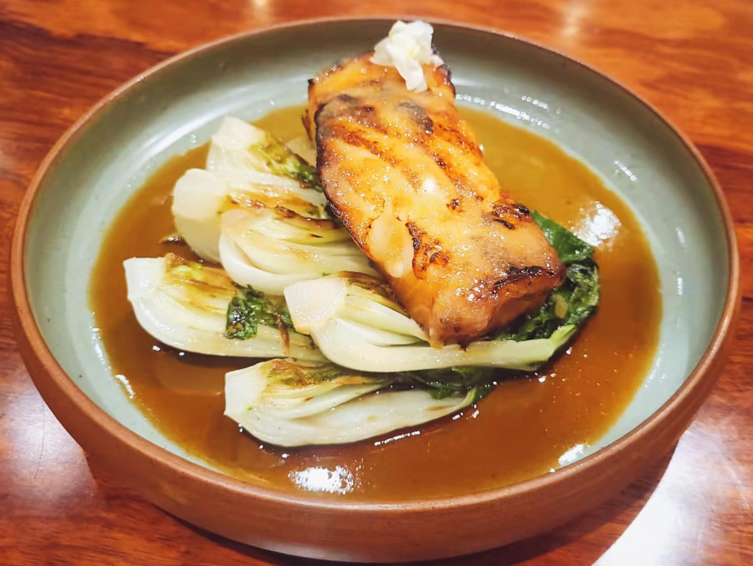 Glossy roasted sea bass fillet in a citron glaze over charred bok choy at a Bay Area restaurant, the kind of scroll-stopping hero dish that makes paid social ads actually work.