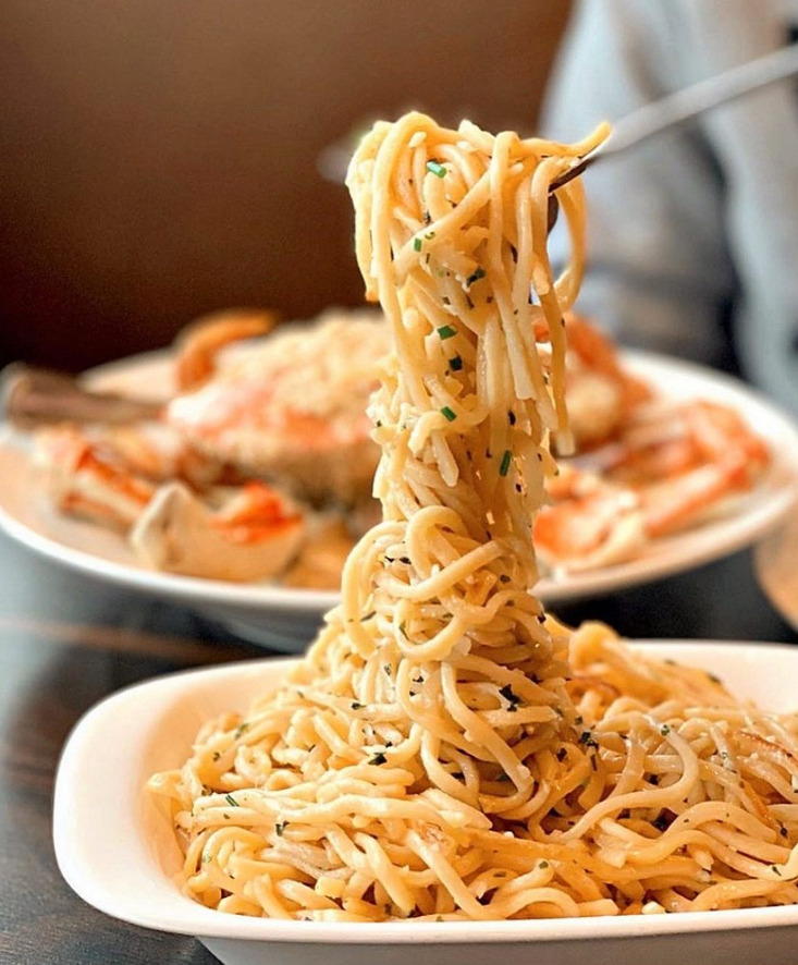 A fork lifting a tall pull of San Francisco style garlic noodles above a white bowl with a garlic crab dish behind it, the kind of tight high contrast dish photo that wins taps on delivery apps for Bay Area restaurants.