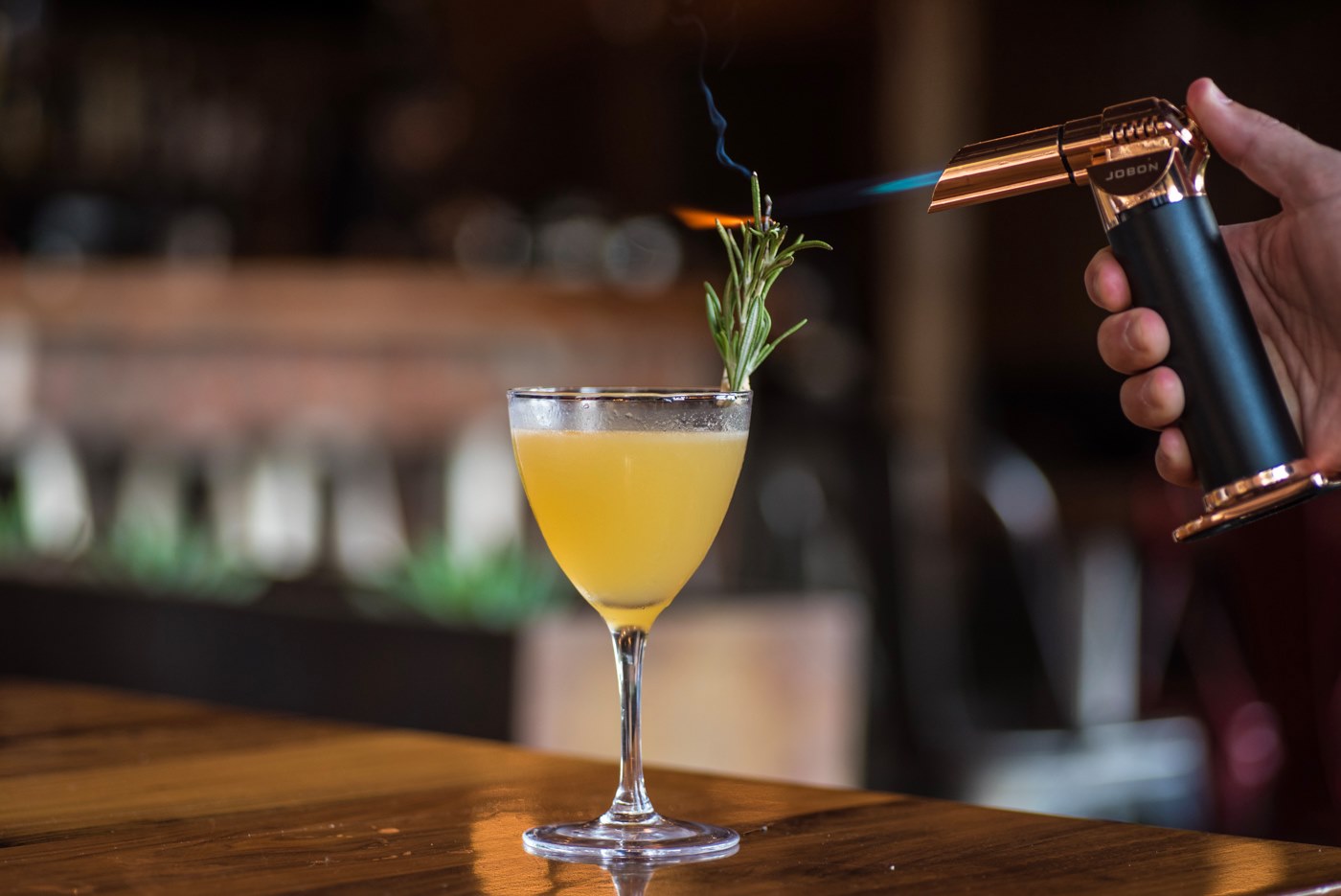 A bartender uses a torch to flame a rosemary sprig garnishing a golden cocktail in a stemmed glass on a warm wooden bar, the kind of made-in-front-of-you moment that makes a Bay Area restaurant’s drinks program easy to market on social media.