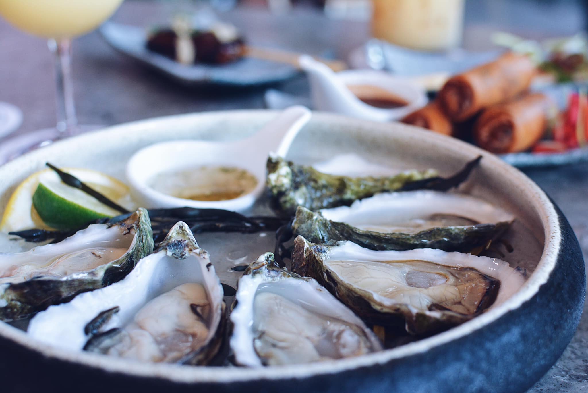 Freshly shucked oysters on the half shell served over ice with lemon and lime at a Bay Area restaurant, with a cocktail and shared plates in the background, the kind of signature dish a new restaurant builds its opening week content around.